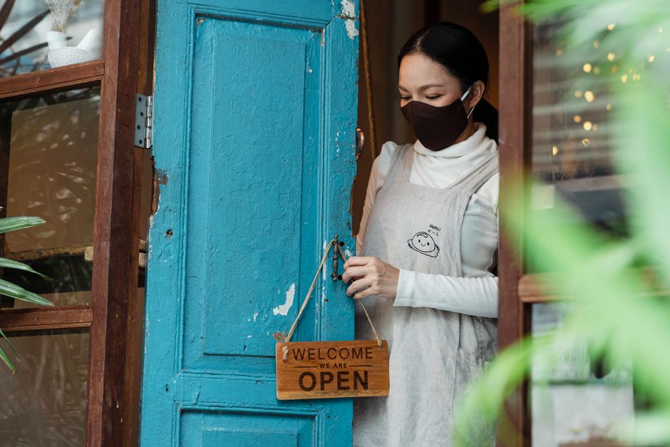Female shopkeeper with face mask setting a welcome sign on a blue door.